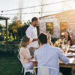 Man giving a speech to group of friends at party. Young people celebrating a special occasion sitting around a table in garden restaurant.